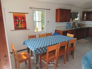 A dining area with a table set for six, wooden chairs, a blue tablecloth, and a view of the kitchen with cabinets and a water dispenser.