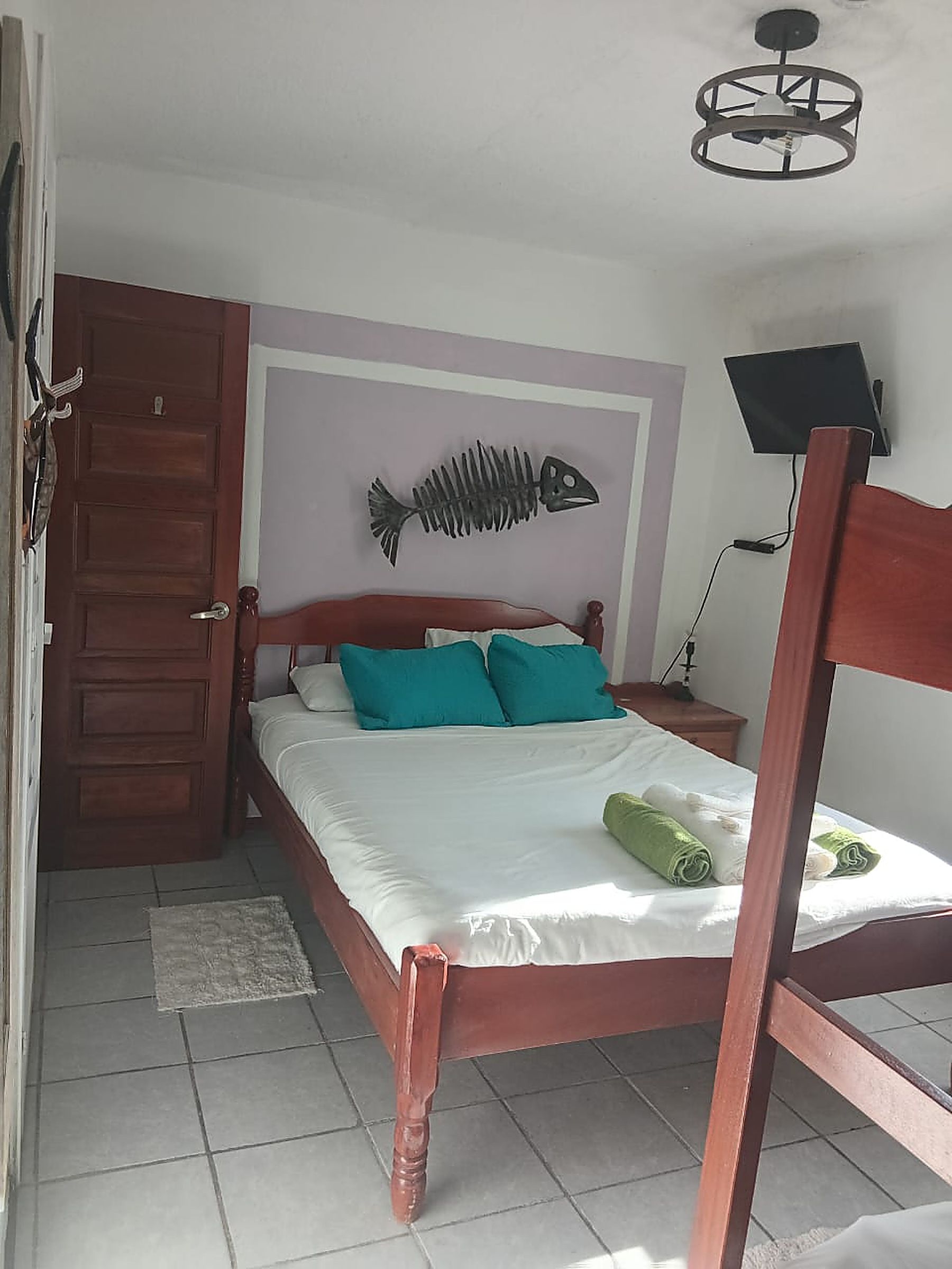A bedroom with a wooden bed, teal pillows, a fish skeleton wall art, a television on a stand, and a door.