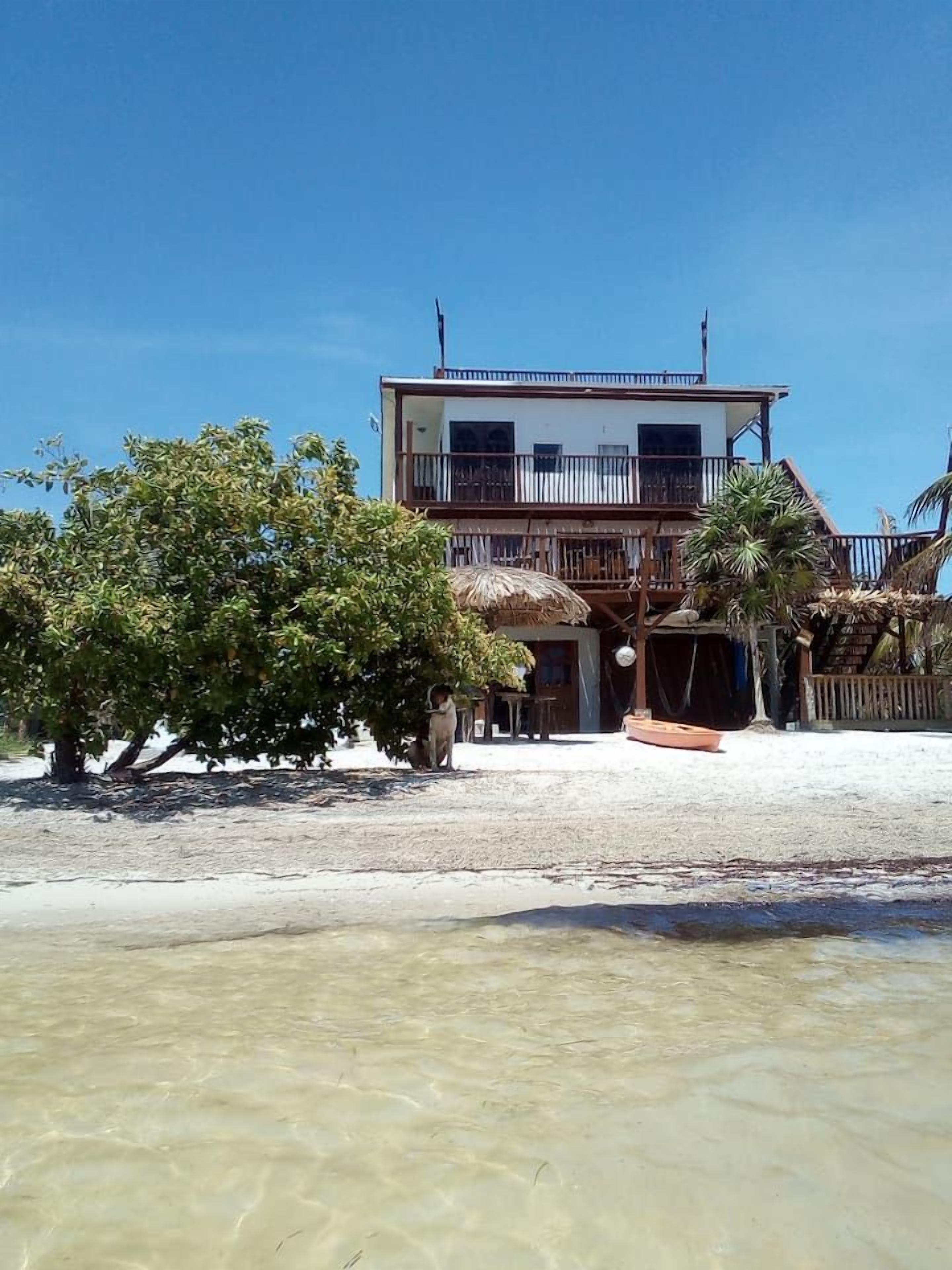 Ein zweistöckiges Gebäude mit Balkon, ein Gebäude mit Strohdach und ein Sandstrand im Vordergrund.