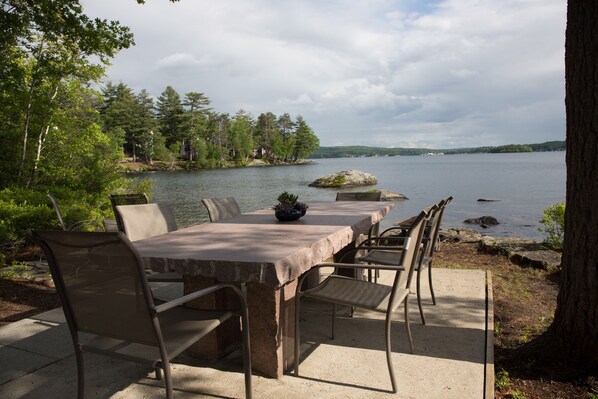 An outdoor dining area with a table and chairs overlooking a lake, surrounded by trees and a rocky shoreline. - RENT YOUR OWN ISLAND on beautiful Lake Winnipesaukee! (Laconia)