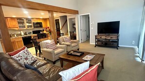 A spacious living room with a brown leather sofa, a wooden coffee table, and a television on a wooden stand. There is a ceiling fan and a kitchen area visible in the background. - Walk 2 Baldy/River, Pool,Hot Tub, Bikes, Private Ski Locker at Mtn, Grt reviews  (Ketchum)