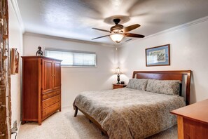 Une chambre à coucher avec un lit, une commode en bois, un ventilateur de plafond, une table de chevet avec une lampe et un tableau encadré accroché au mur.