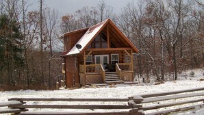A wooden cabin with a porch and steps leading to the entrance, surrounded by snow and trees.