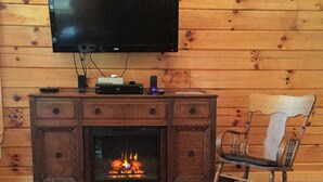 A wooden cabin interior with a television mounted on the wall, a fireplace with a visible fire, a wooden cabinet with electronic devices, and a rocking chair.