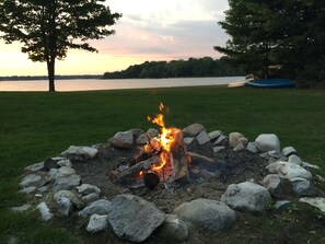 Ein Lagerfeuer, umringt von Steinen, in einer grünen Landschaft mit einem See und Bäumen im Hintergrund.