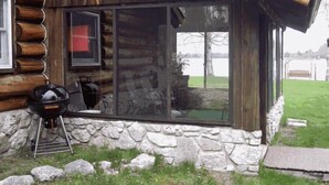 Une cabane en rondins avec des fondations en pierre, une grande fenêtre et un barbecue à l’extérieur.