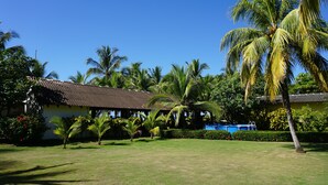 A well-maintained garden with palm trees, a small building with a tiled roof, and a clear blue sky.