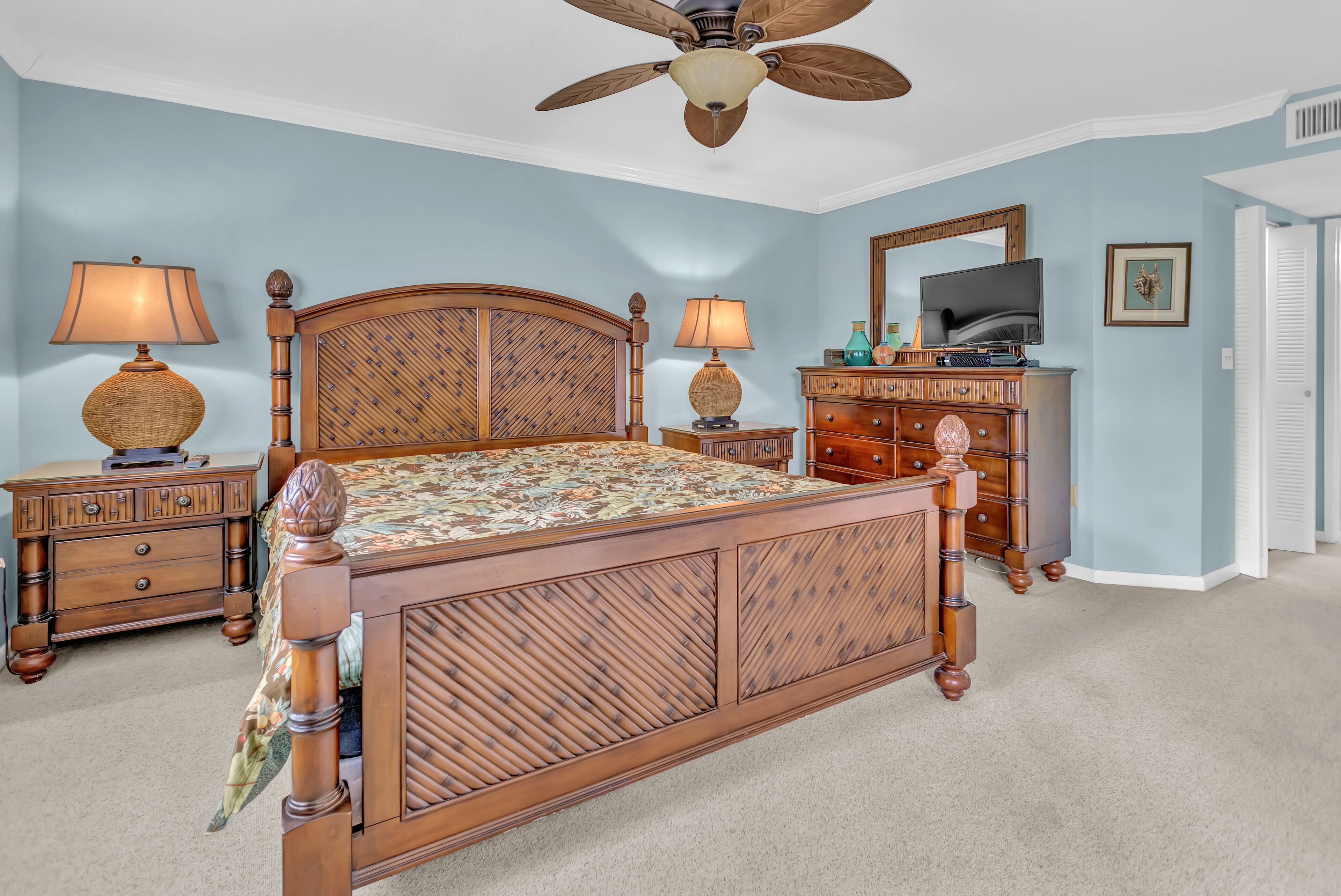 Une chambre à coucher avec un lit en bois, deux tables de chevet, une commode, un ventilateur de plafond, un téléviseur et une porte.