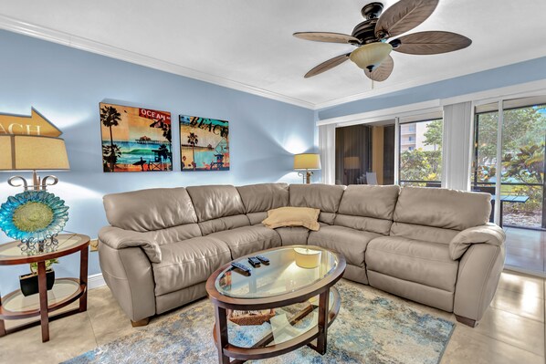 A living room with a beige sectional sofa, a glass coffee table, and a ceiling fan. There are two wall art pieces depicting beach scenes and a decorative lamp on a side table. - Comfy Condo at Jupiter Bay Resort, with easy beach access (Jupiter)