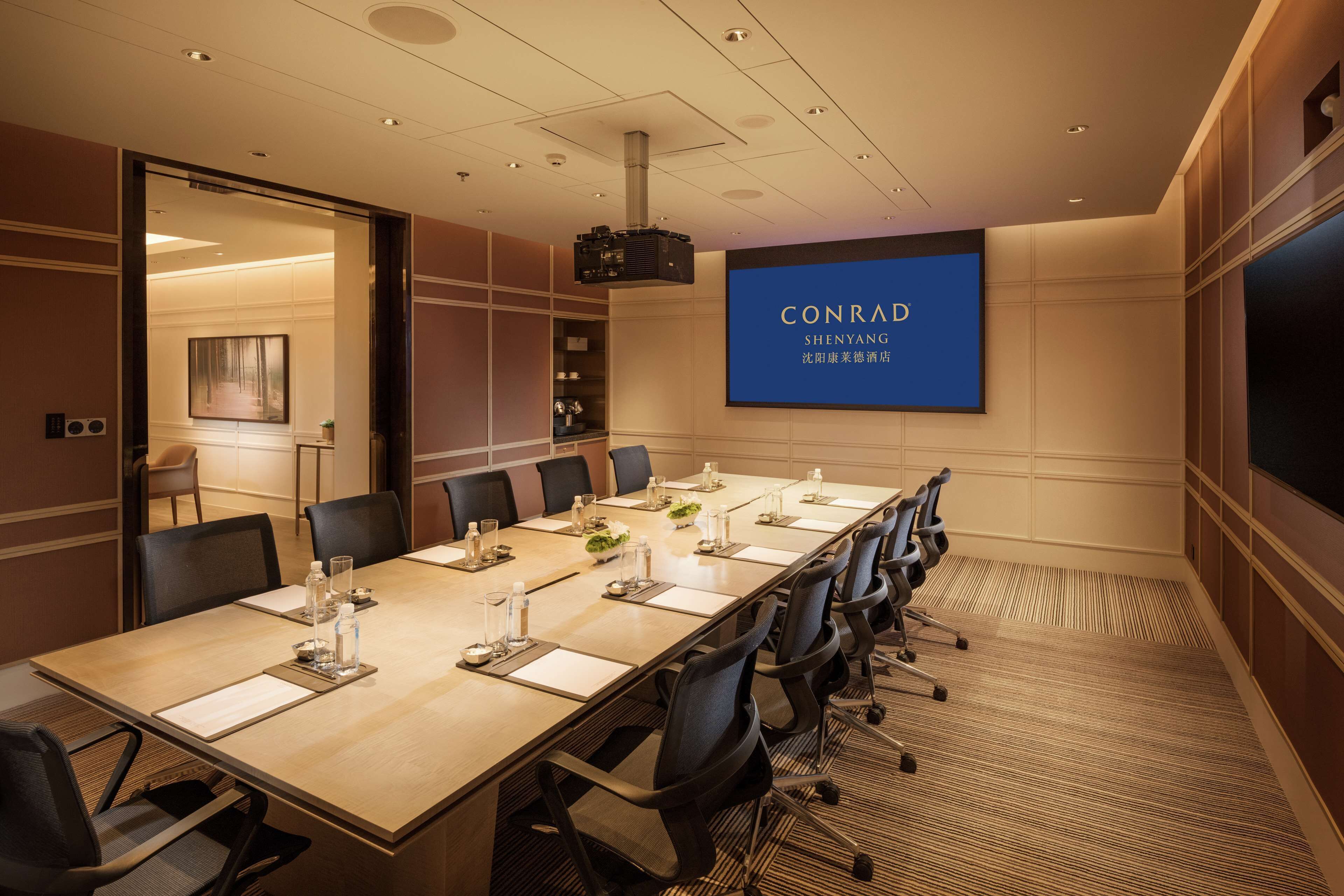 A conference room with a long table, chairs, a projector screen, and a large blue sign with "CONRAD SHENYANG" text.