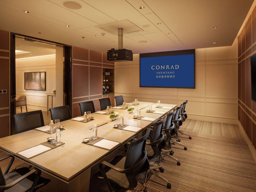 A conference room with a long table, chairs, a projector screen, and a large blue sign with "CONRAD SHENYANG" text.