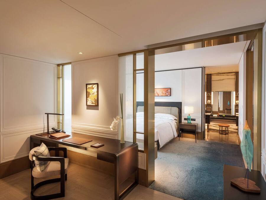 Quarto de hotel com escrivaninha, cadeira, cama e banheiro visível por uma porta aberta.