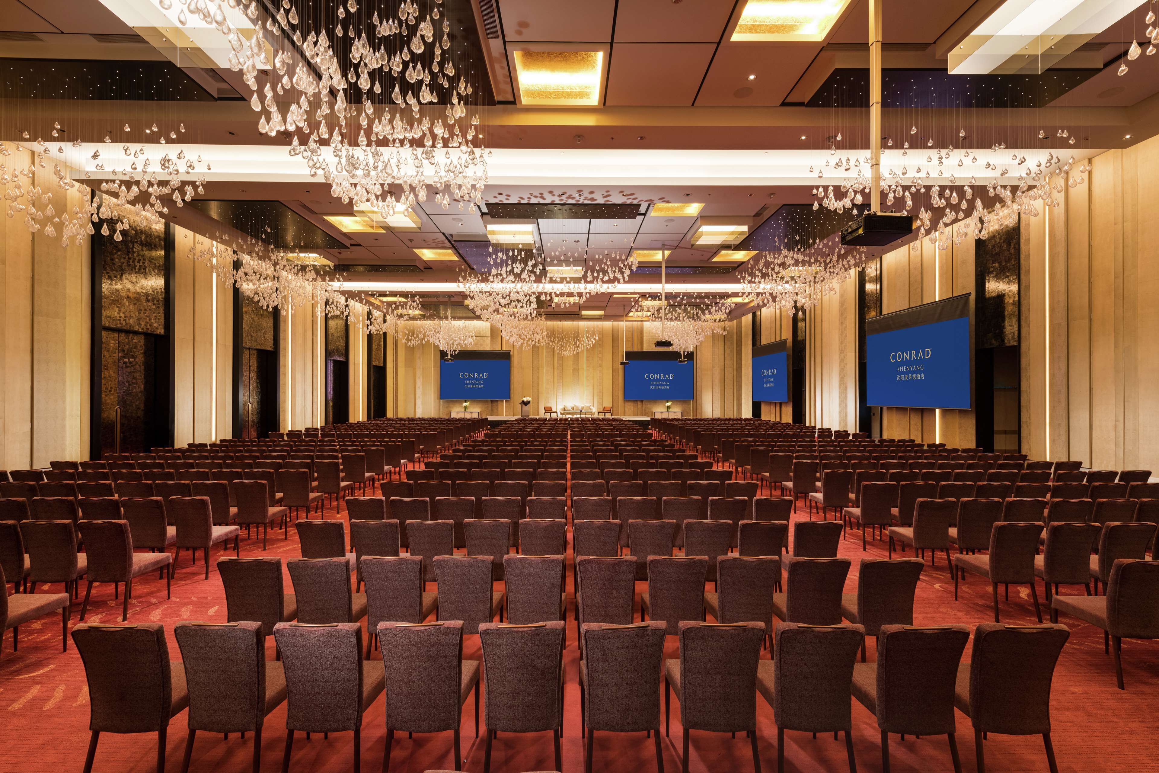 Un salón de conferencias con filas de sillas, un escenario y pantallas mostrando la marca 'CONRAD'.