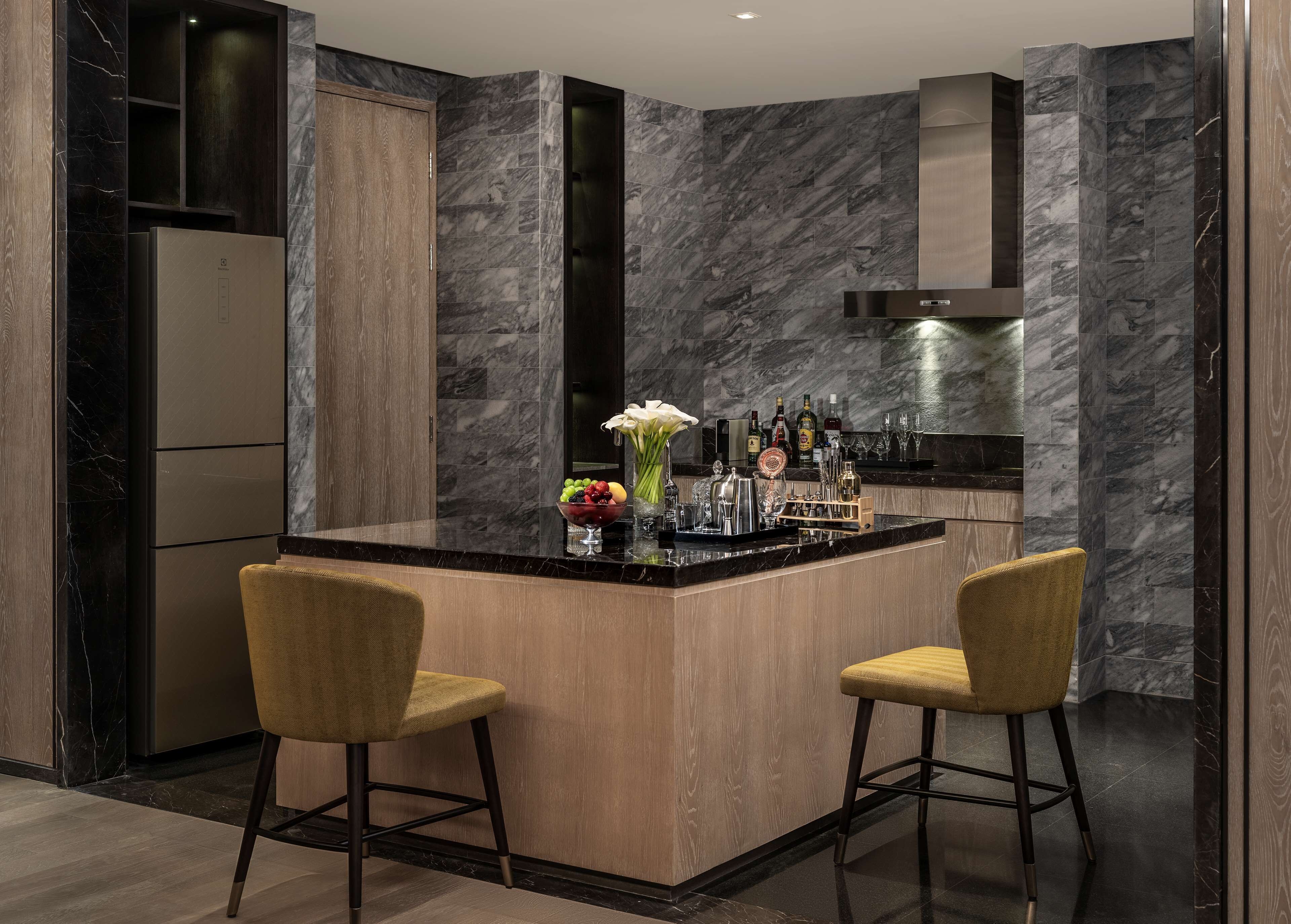 A modern kitchen with marble walls, a central island with bar stools, a built-in refrigerator, and a variety of barware.