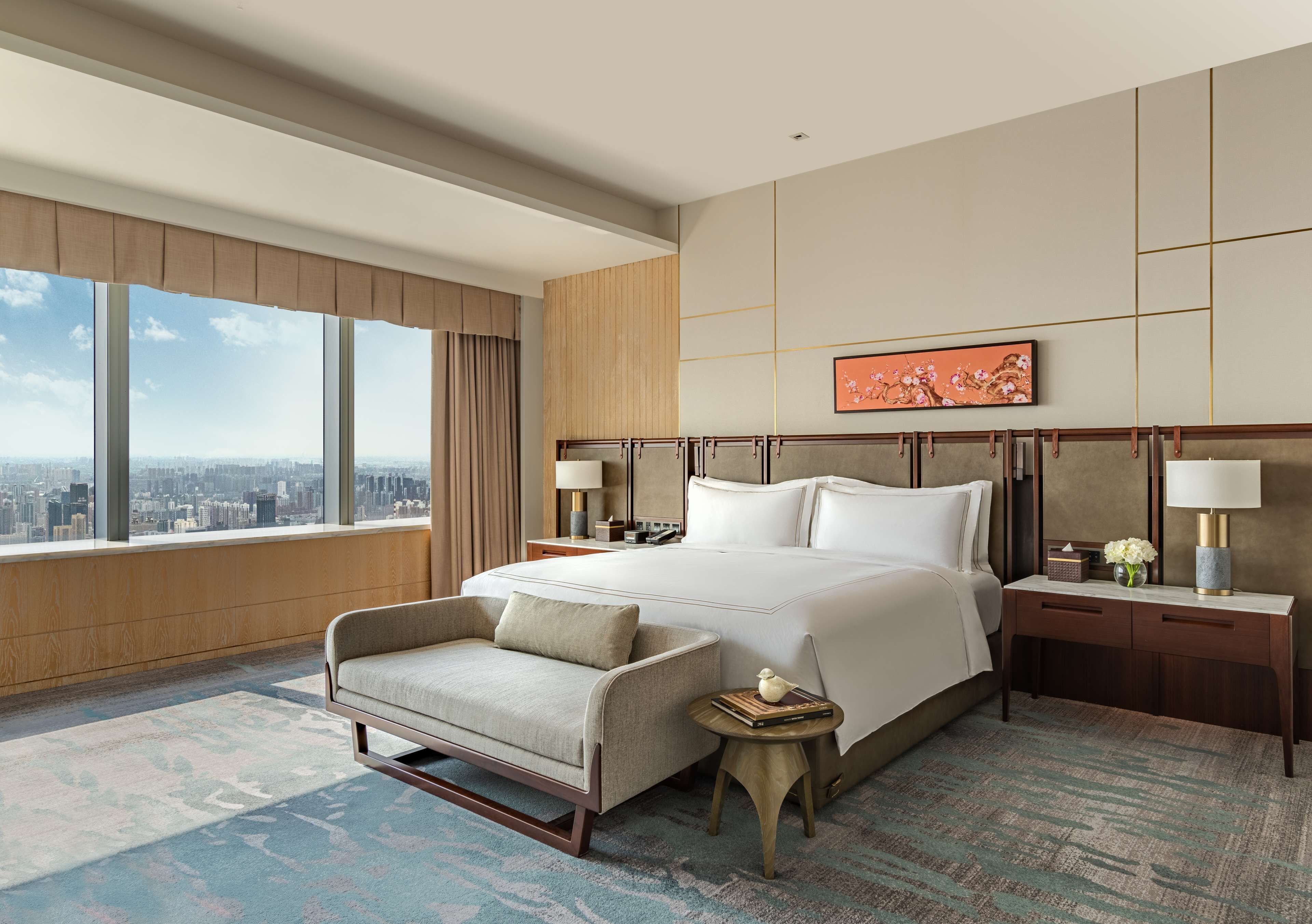 A hotel room with a large bed, a sofa, a small table, and a view of the city through large windows.