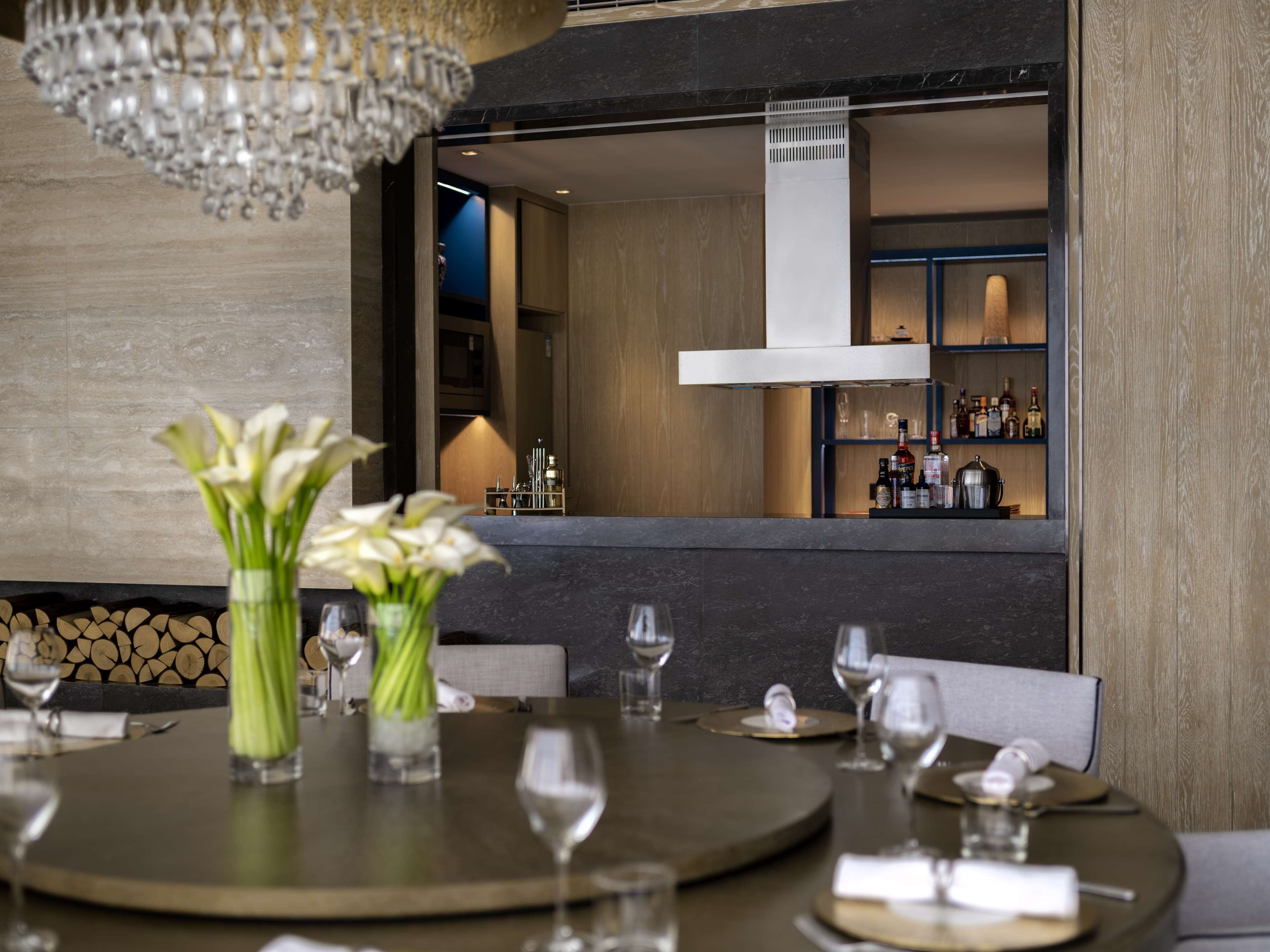 Un comedor con una mesa redonda dispuesta para dos, un florero con flores y una vista del área de bar al fondo.