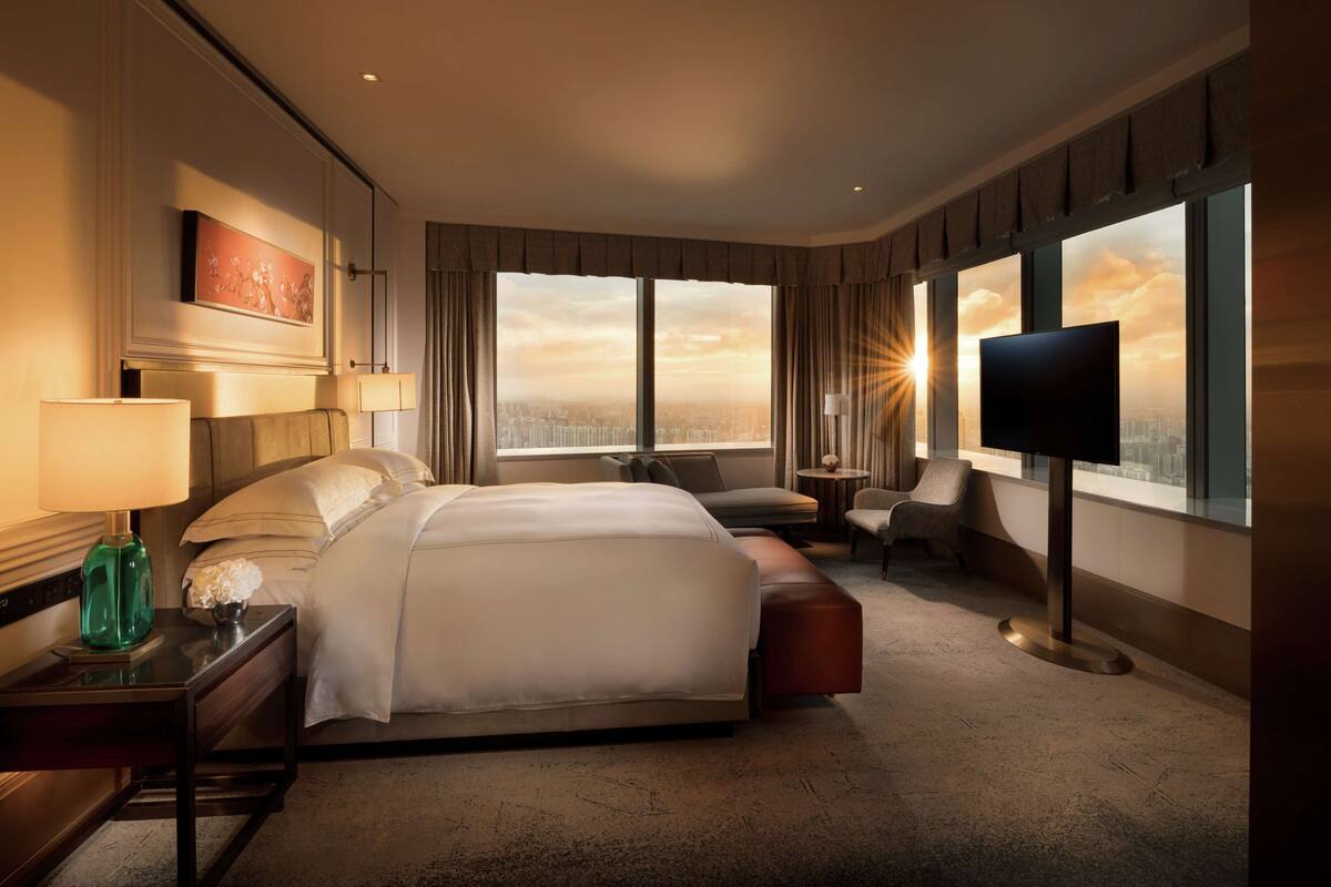 A hotel room with a large bed, a desk with a lamp, a TV, and a view of the cityscape.