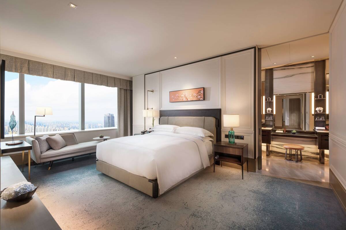 A modern hotel room with a large bed, a sofa, a desk, and a view of the city.