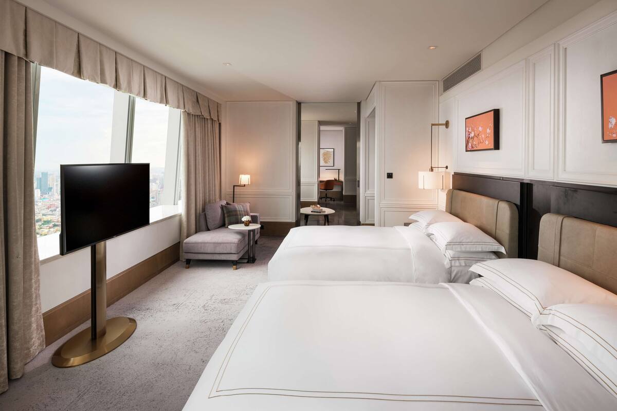 A hotel room with two beds, a TV, a desk, and a view of the city.