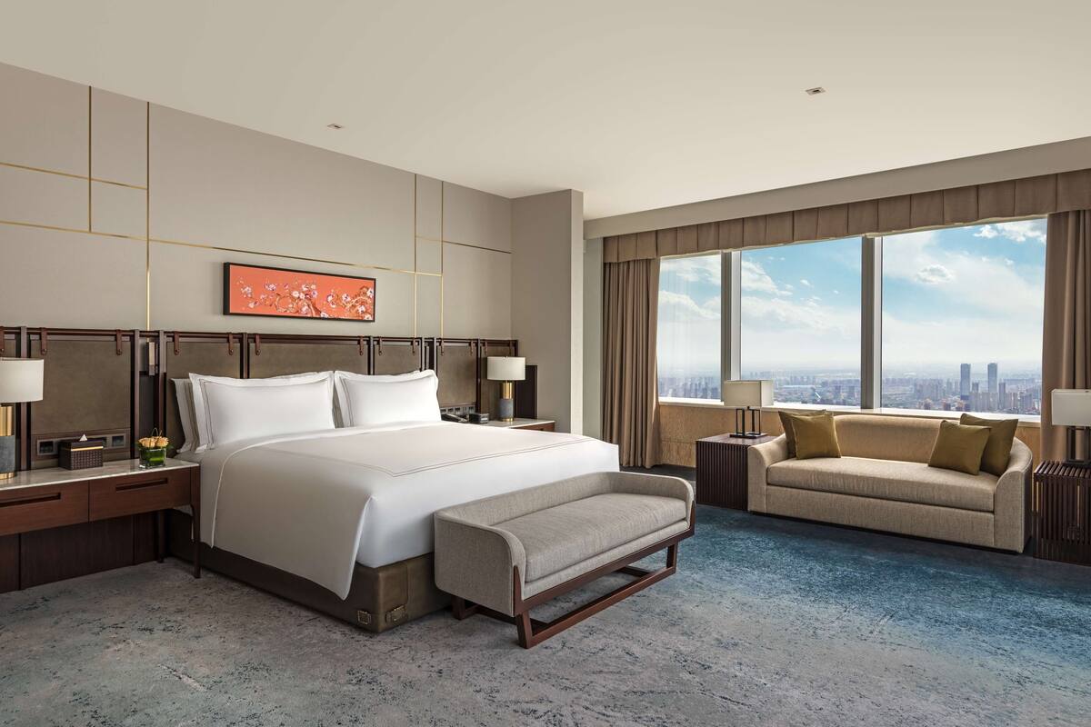 A modern hotel room with a large bed, a sofa, a city view, and a decorative painting.