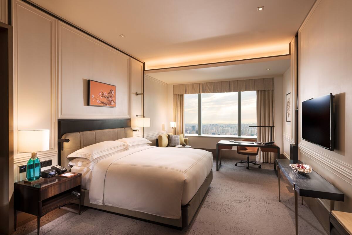 A hotel room with a large bed, a desk, a chair, a TV, and a window with a city view.