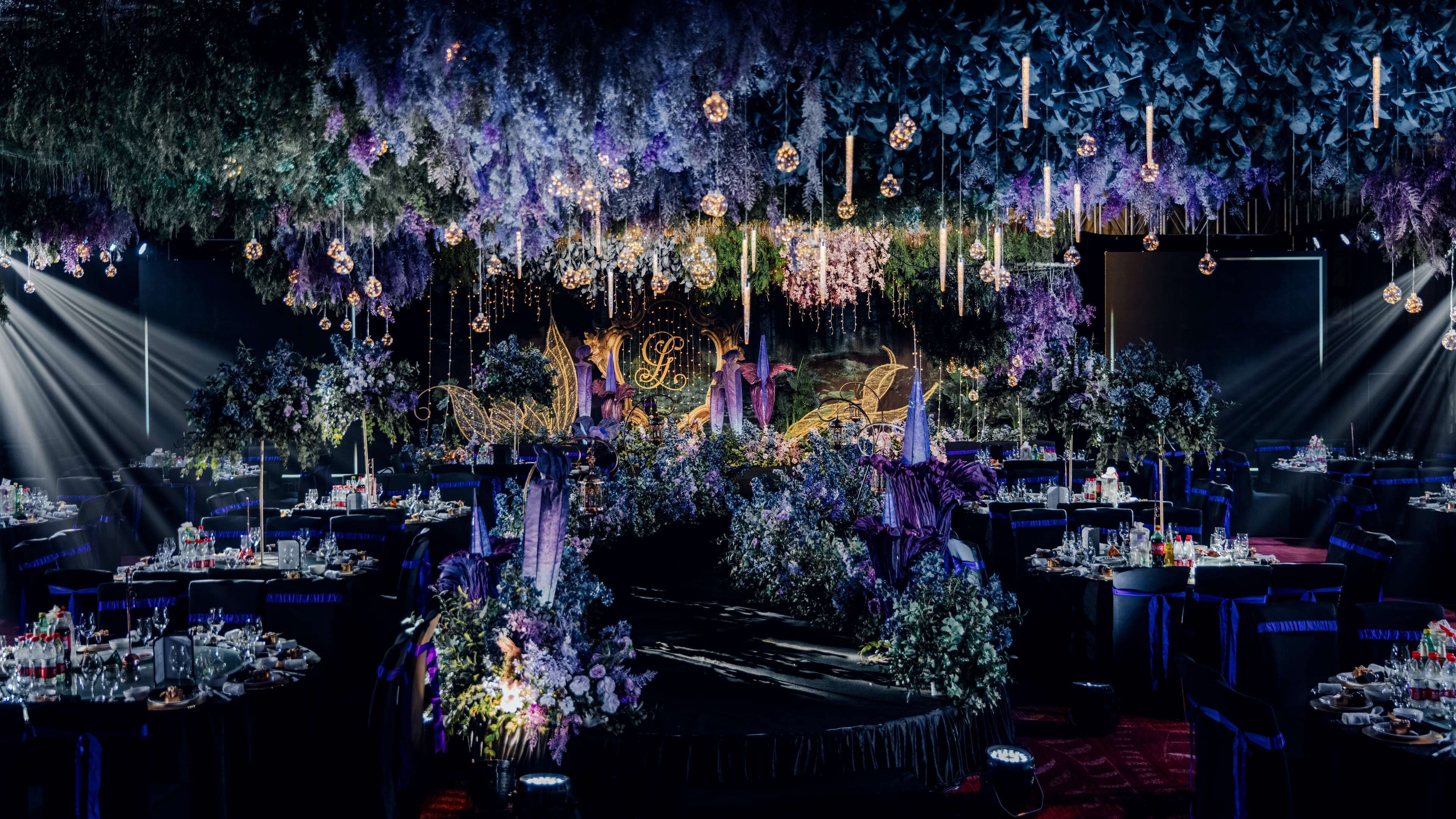 A formal event space with tables set for dining, decorated with floral arrangements and hanging lights.