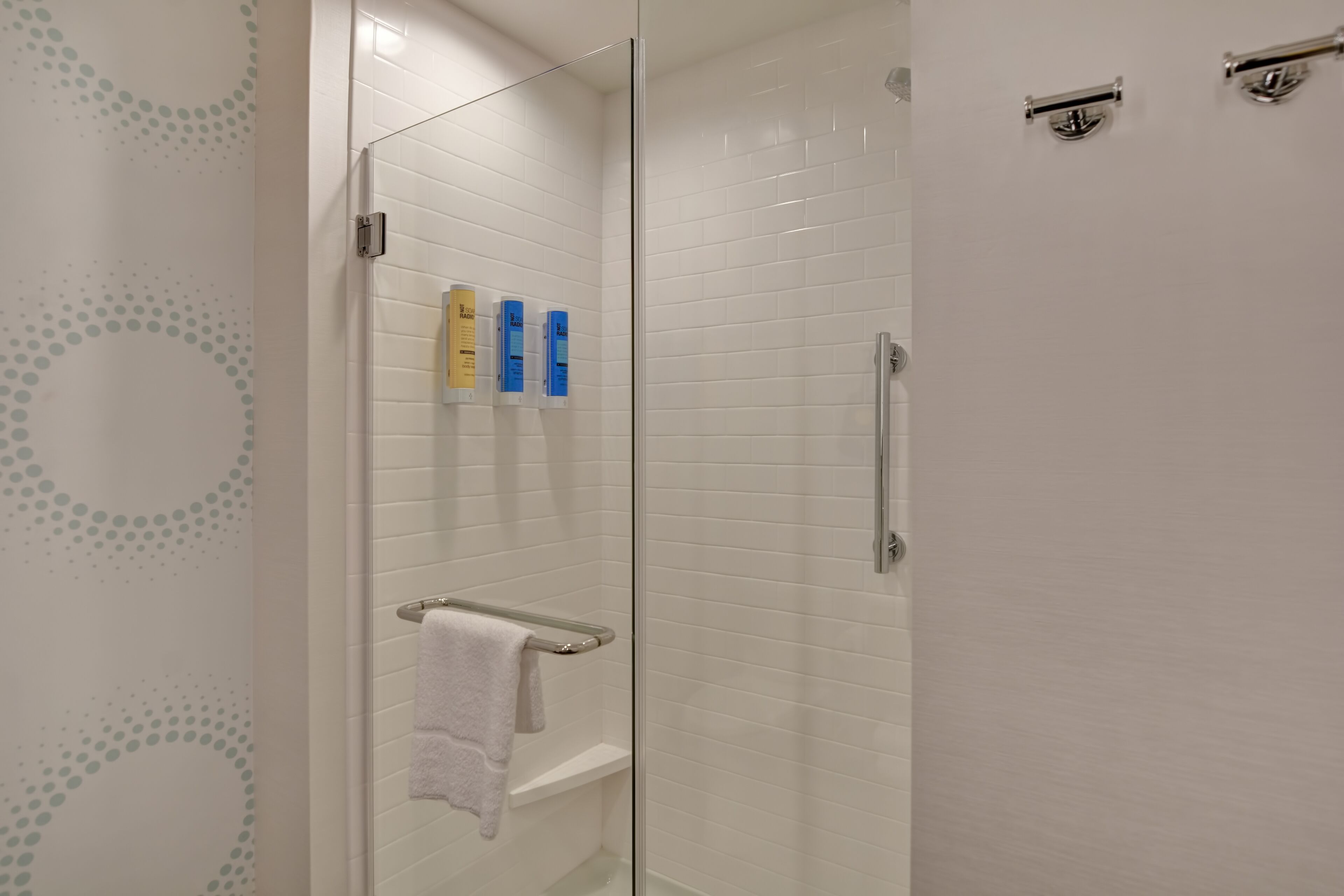 A modern bathroom with white tiled walls, a glass shower enclosure, a towel rack, and two toiletries on the wall.