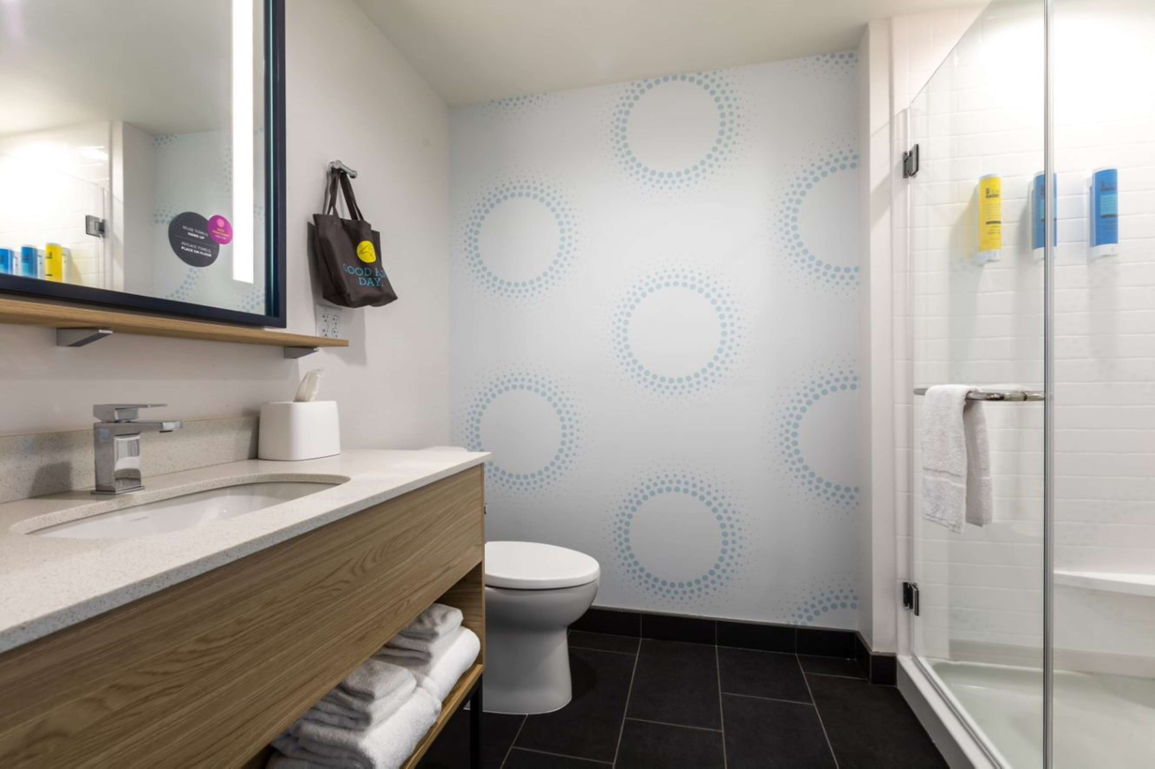 Baño con diseño moderno, que incluye un mueble de tocador de madera, un lavamanos blanco, un inodoro y una cabina de ducha con mampara de vidrio.
