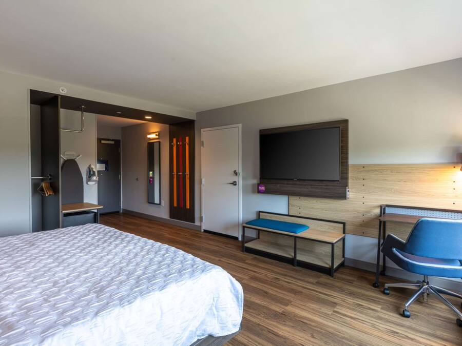 A hotel room with a bed, a desk with a chair, a TV mounted on the wall, and a bench.