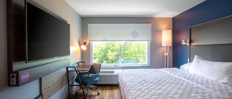 A hotel room with a large bed, a desk with a chair, a flat-screen TV, a window with a view of trees, and a headboard.