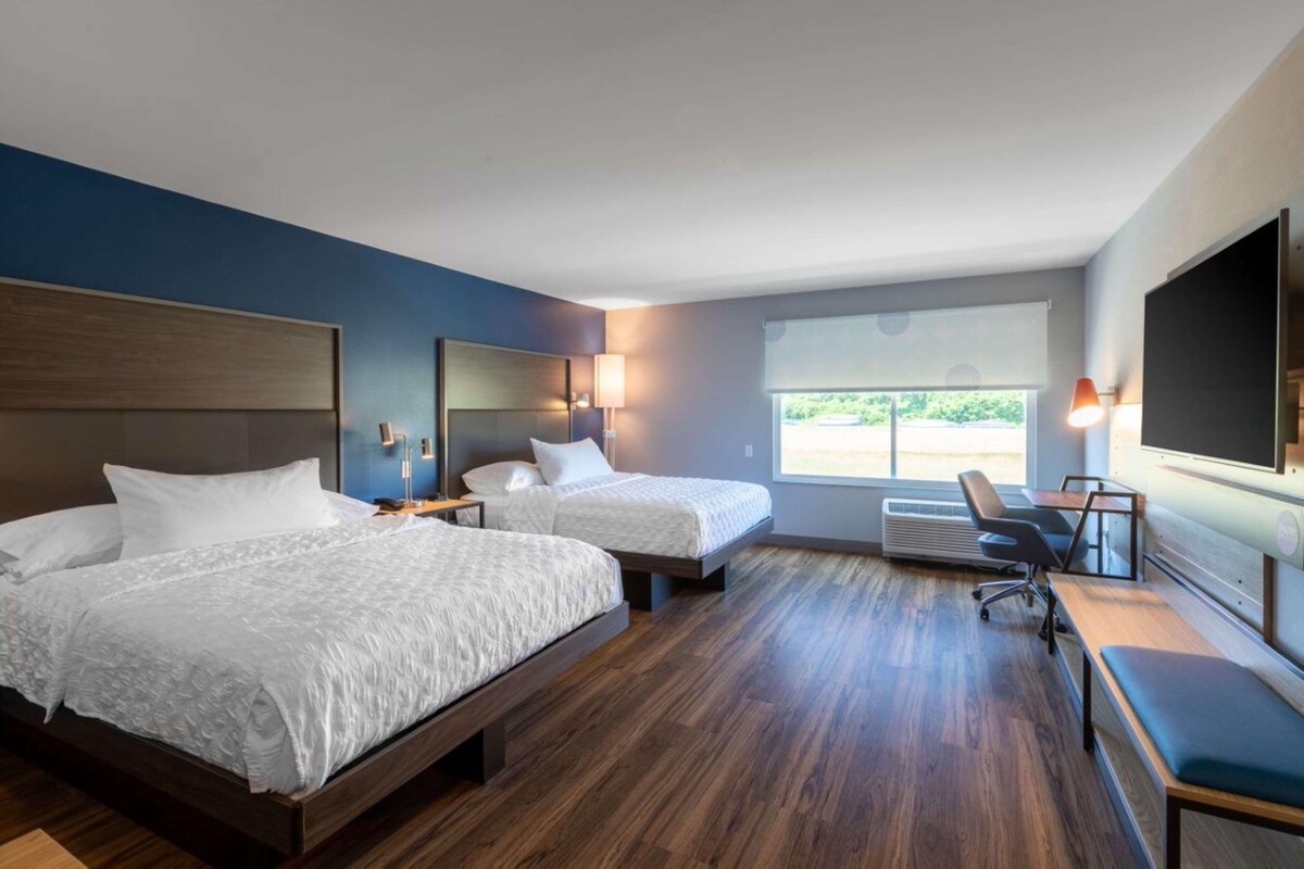 A hotel room with two beds, a desk, a chair, a TV, and a large window.