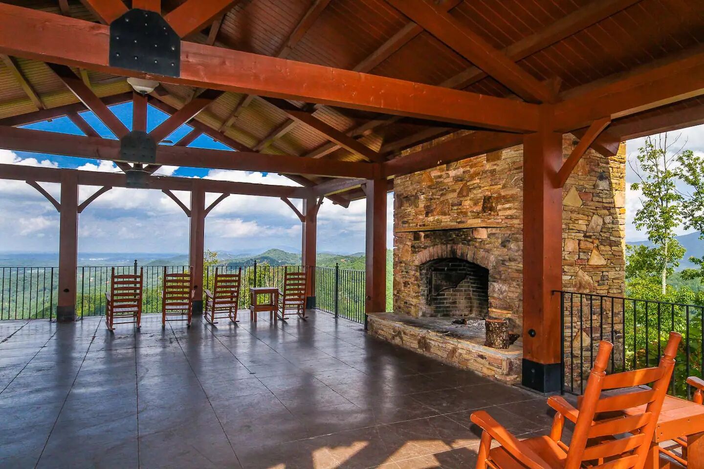 A covered outdoor area with a stone fireplace, wooden chairs, and a view of the landscape.