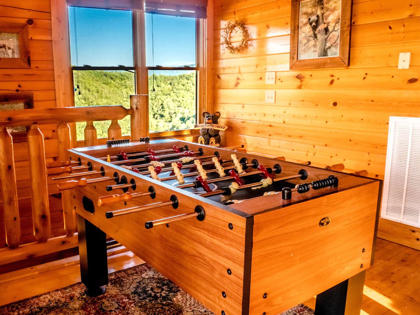 Una habitación con piso de madera, una mesa de fútbol de mesa, una ventana con vistas a colinas verdes y un cuadro en la pared.