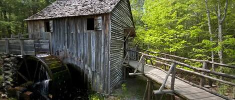 A rustic wooden structure with a water wheel, surrounded by lush greenery and a wooden bridge.