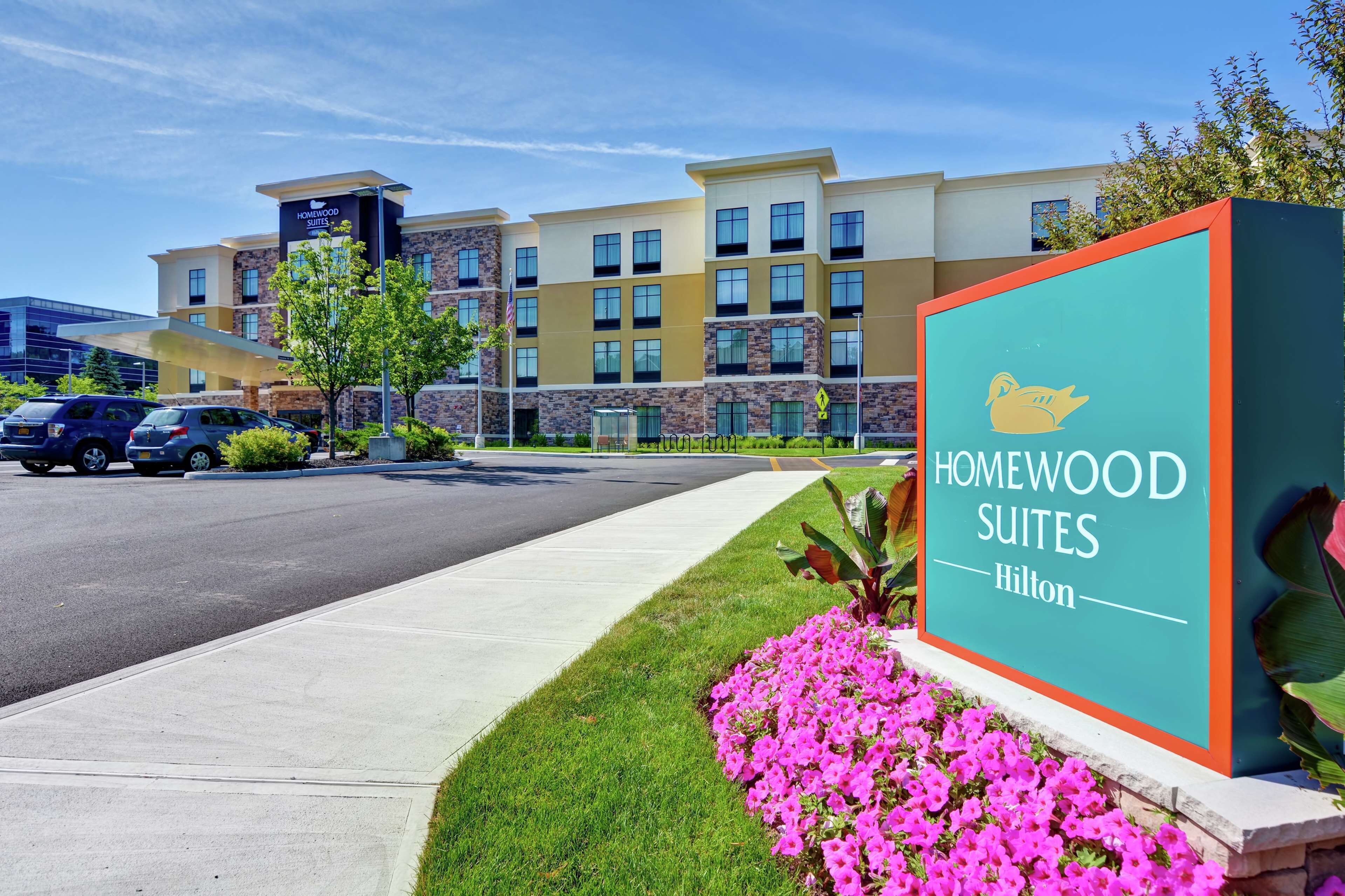 A hotel entrance with a sign reading "Homewood Suites Hilton" and a parking lot with cars.