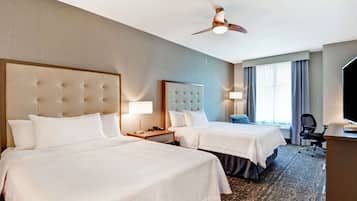 Quarto de hotel com duas camas, uma escrivaninha, uma cadeira, ventoinha no teto e janela com cortinas.