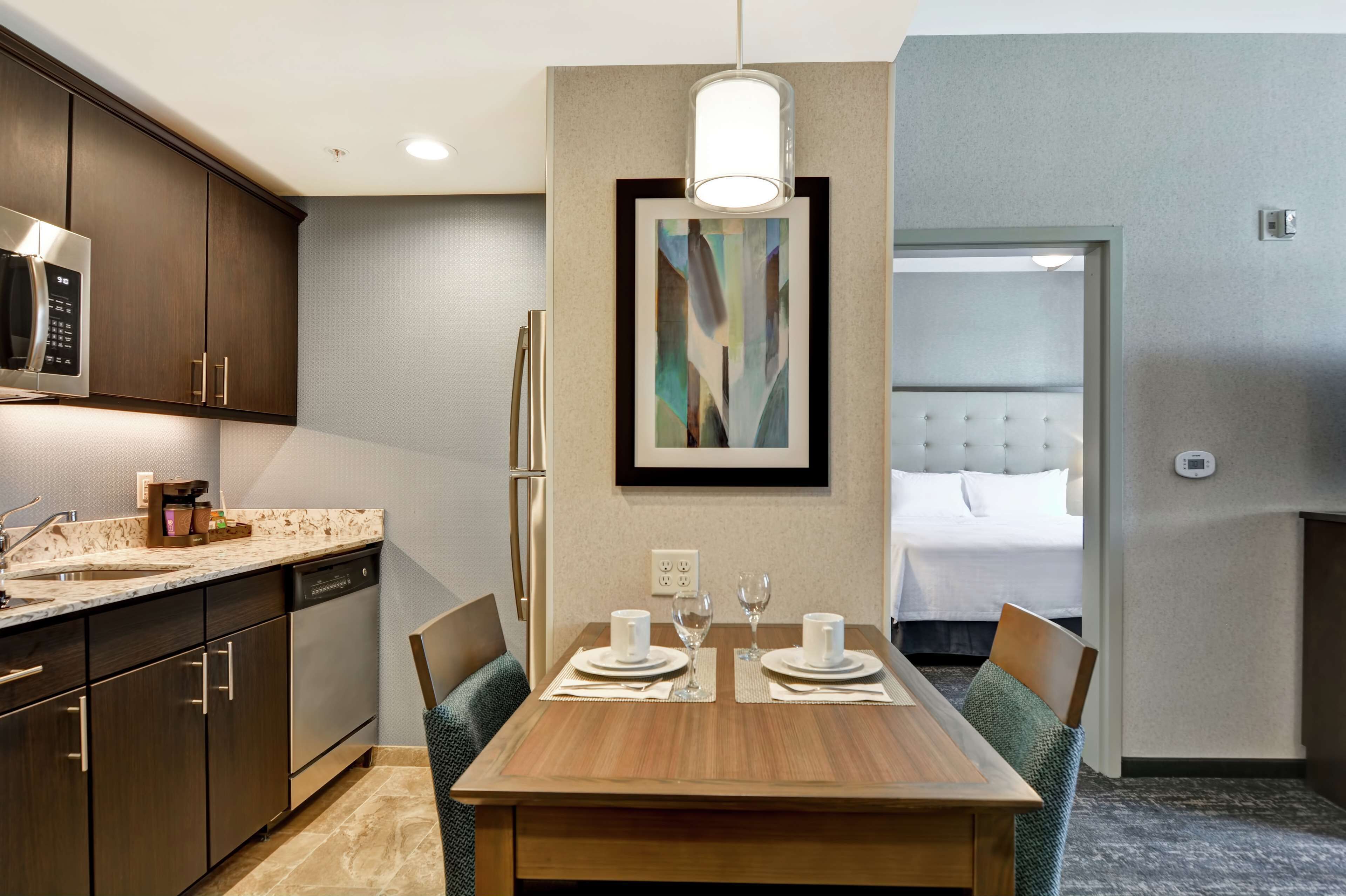 A modern hotel room with a dining area, kitchenette, and a bed with a tufted headboard.