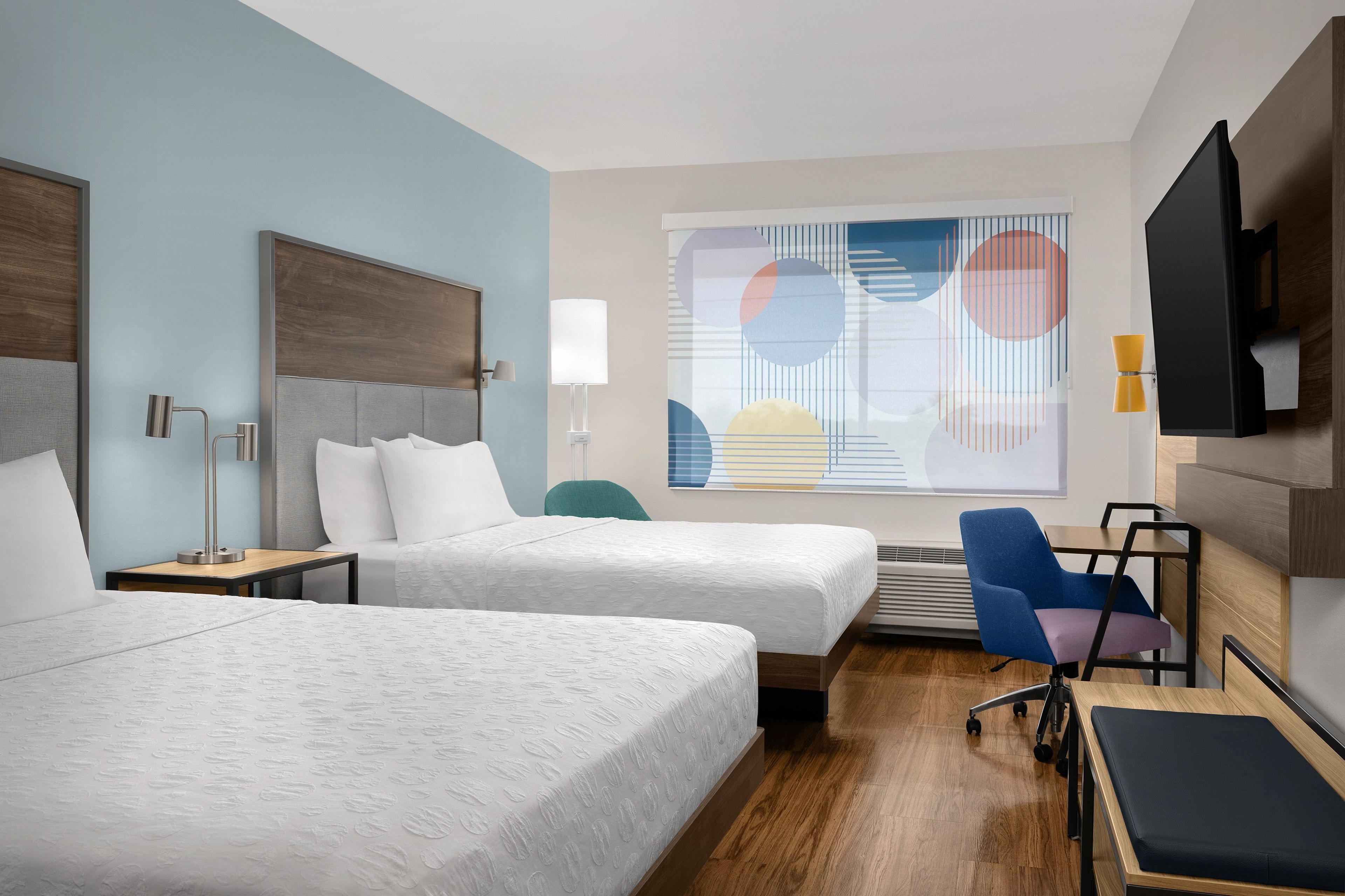 Habitación de hotel con dos camas, un escritorio, una silla, un televisor y un cuadro abstracto de grandes dimensiones en la pared.