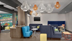 A modern hotel lobby with a reception desk, blue and gray seating, a TV screen, and unique pendant lighting. - Tru by Hilton Orangeburg (Orangeburg)
