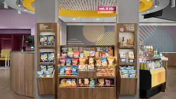 Een hapjesdisplay met verschillende chips en snacks, een koffieapparaat en een bord dat een 24/7 winkel aangeeft.