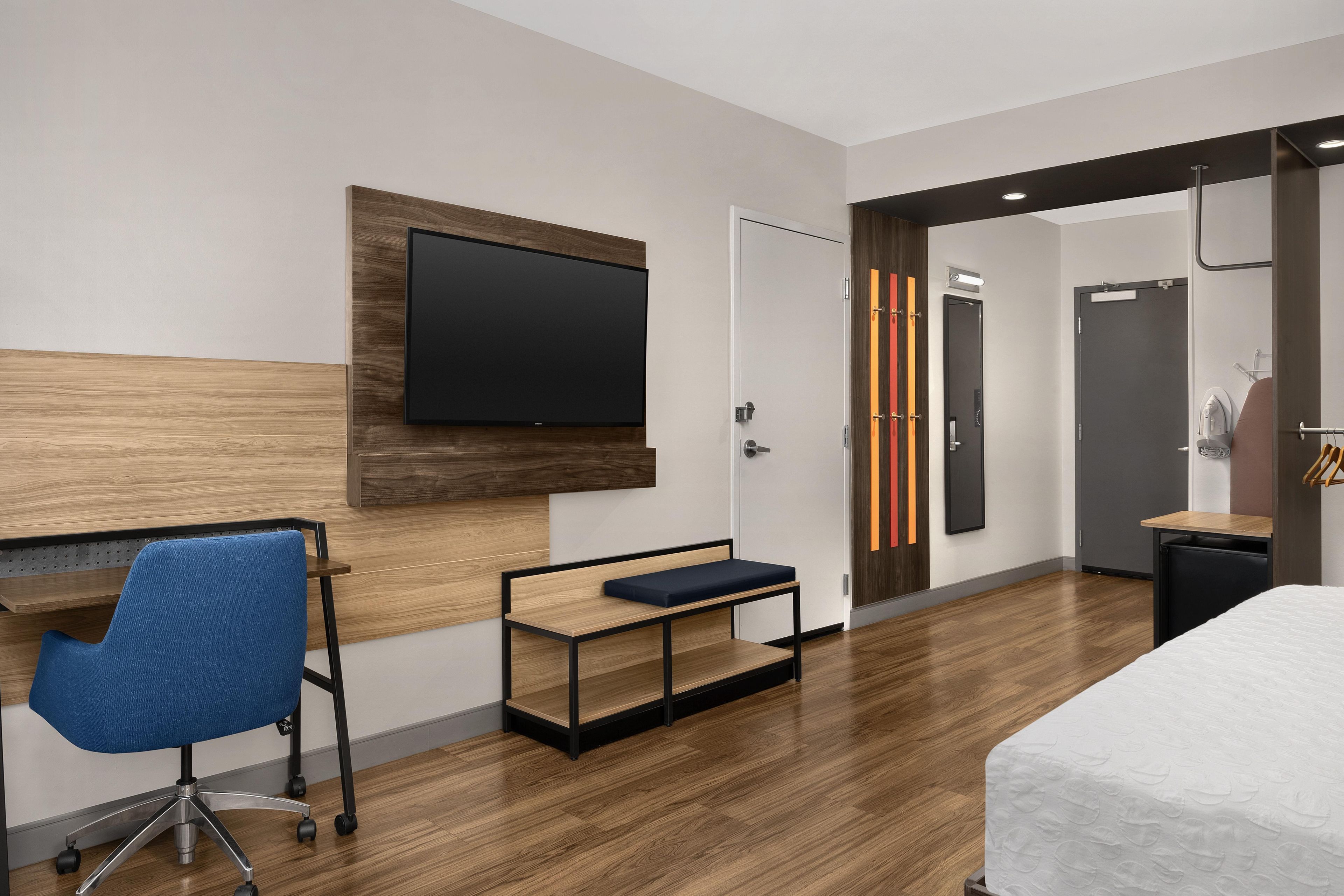A modern hotel room with a flat-screen TV, a desk with a chair, a bed, and a wardrobe.