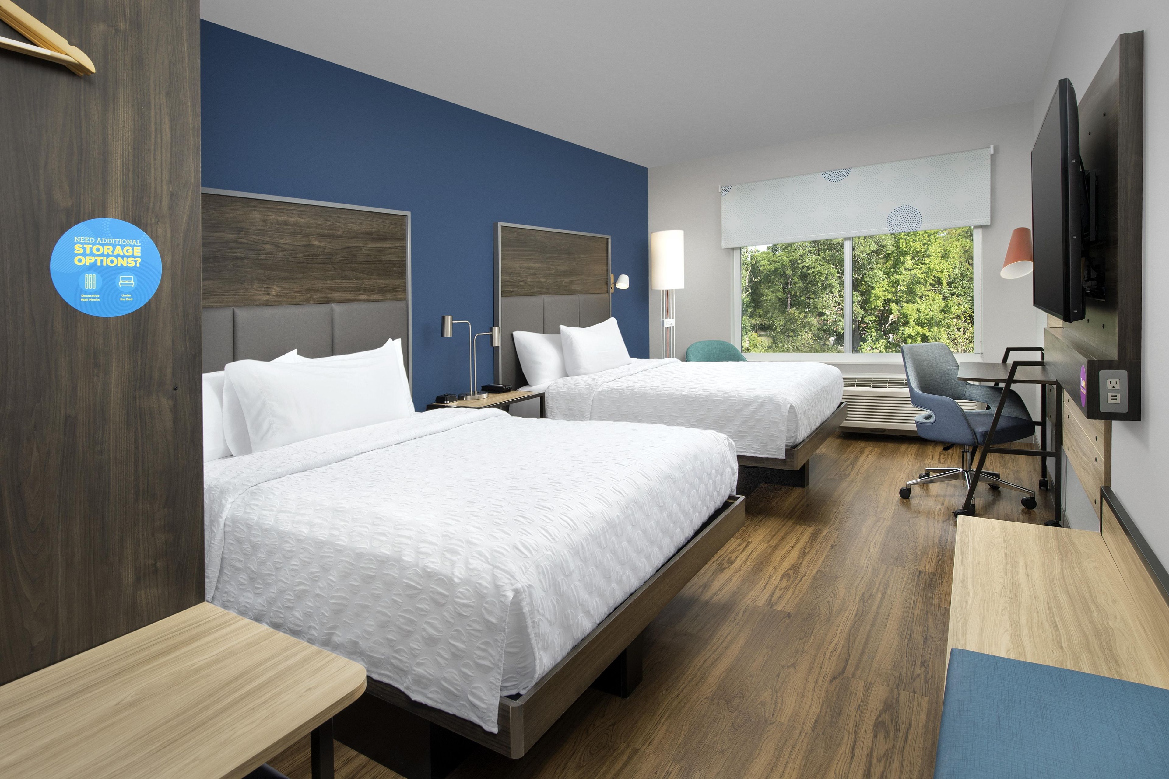 A hotel room with two beds, a desk, a chair, a TV, and a window with a view of trees.