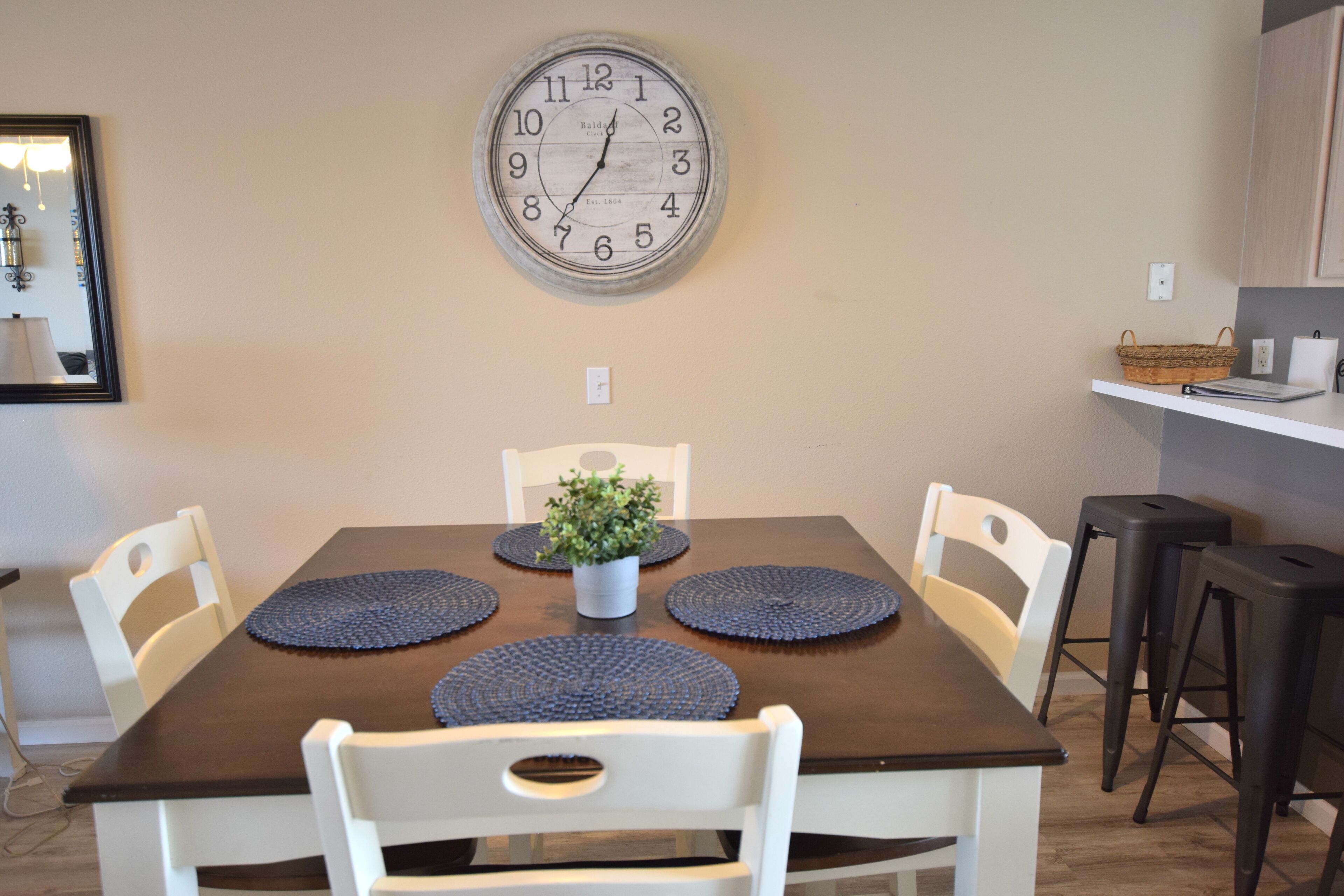 A dining area with a wooden table, white chairs, a large wall clock, and a potted plant.