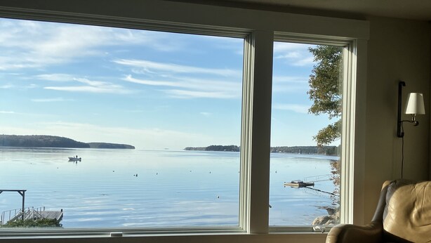 A room with a large window overlooking a calm lake, a chair, and a small stool. - Spectacular Waterfront Retreat Close To Portland!   (West Bath)