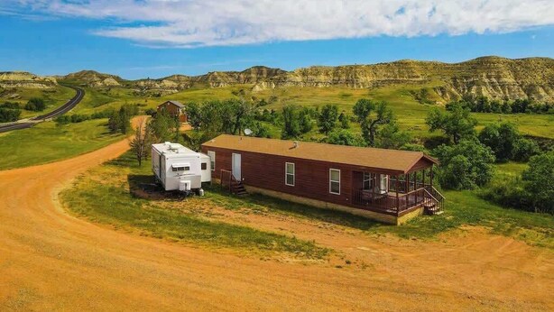 A single-story RV with a porch, a small attached building, and a dirt road leading to a highway.