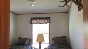 A bedroom with two beds, a window with a lace curtain, a lamp on a nightstand, and a deer skull on the wall.