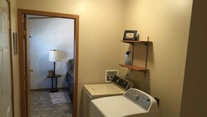 A laundry room with two washing machines, a small table with a lamp, and a shelf with a picture frame and a bottle.