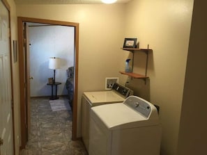 A laundry room with two washing machines, a small table with a lamp, and a shelf with a picture frame and a bottle.