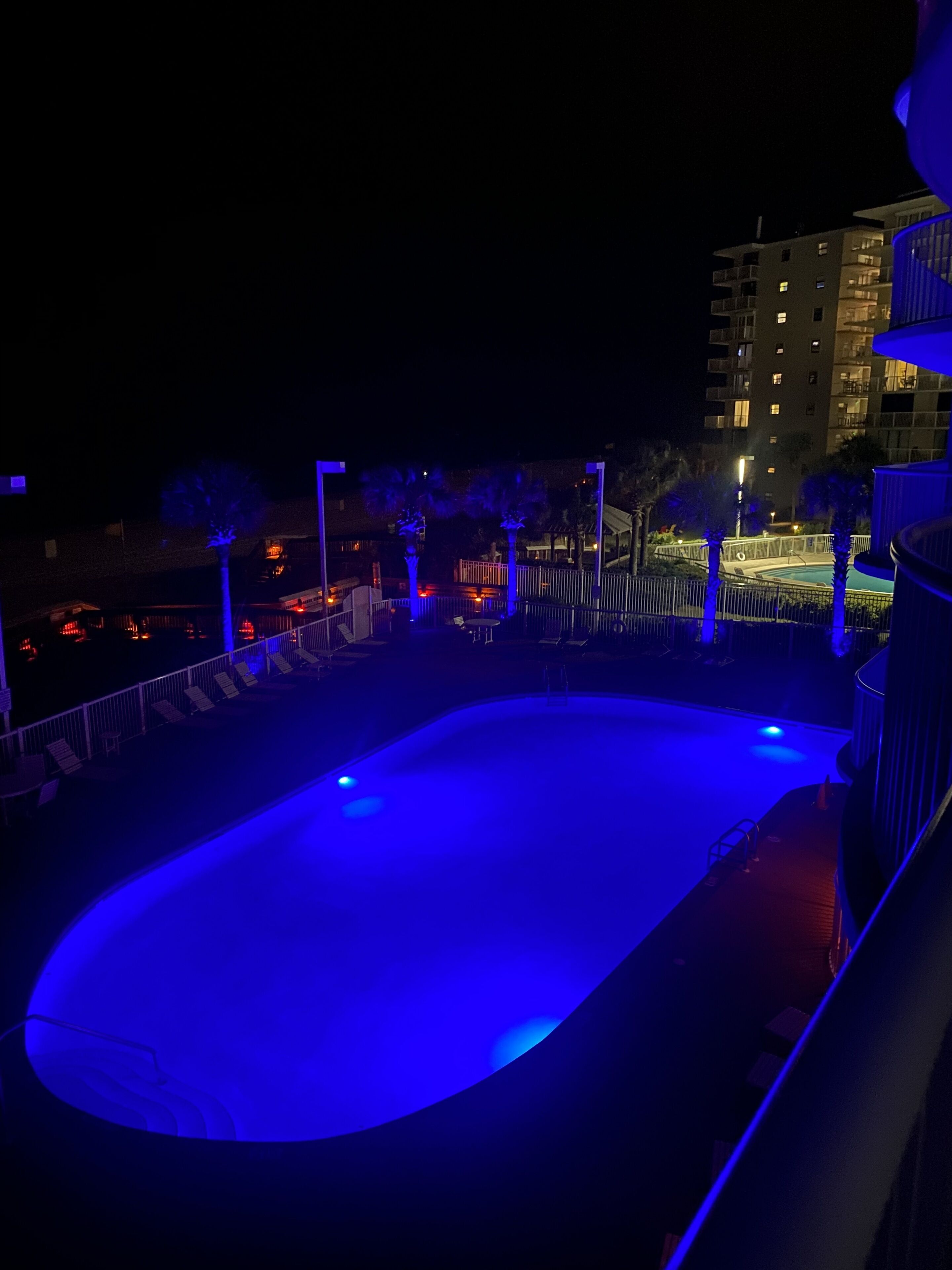 Vista nocturna de una piscina iluminada con luz azul, rodeada por una valla y palmeras, con edificios al fondo.