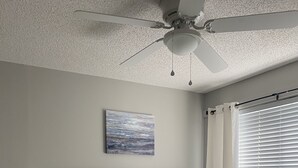 Quarto com cama, mesa de cabeceira, abajur e ventilador de teto.